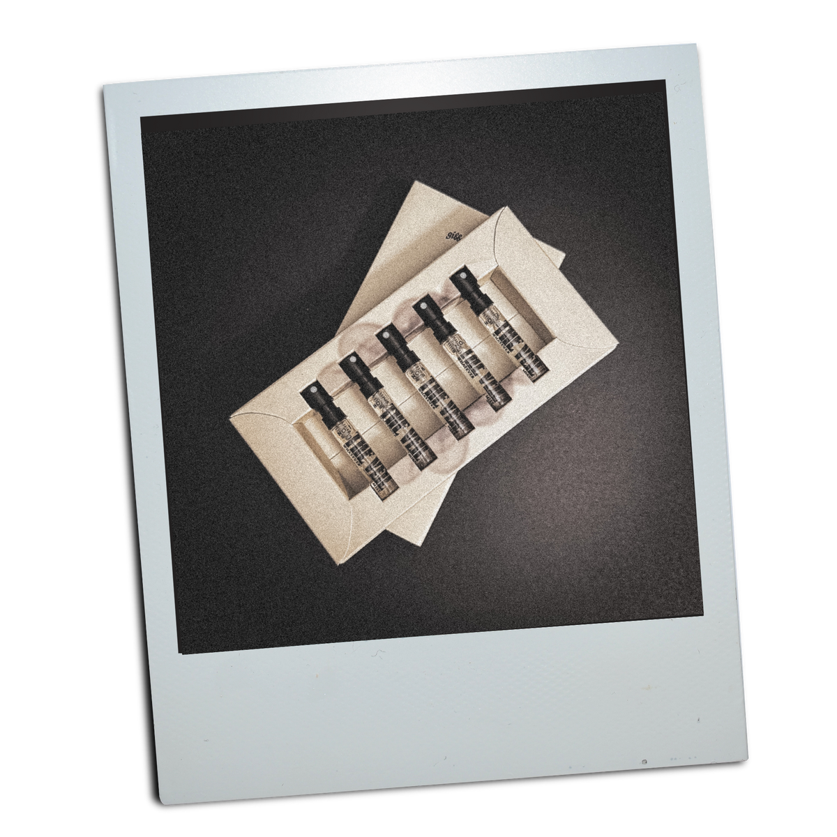 Polaroid-style photo of small perfume bottles with ‘THE VEIL’ on them and a display box for them on a grey background